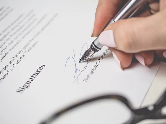 Hand signing the signature line of a printed contract with a fountain pen beside glasses