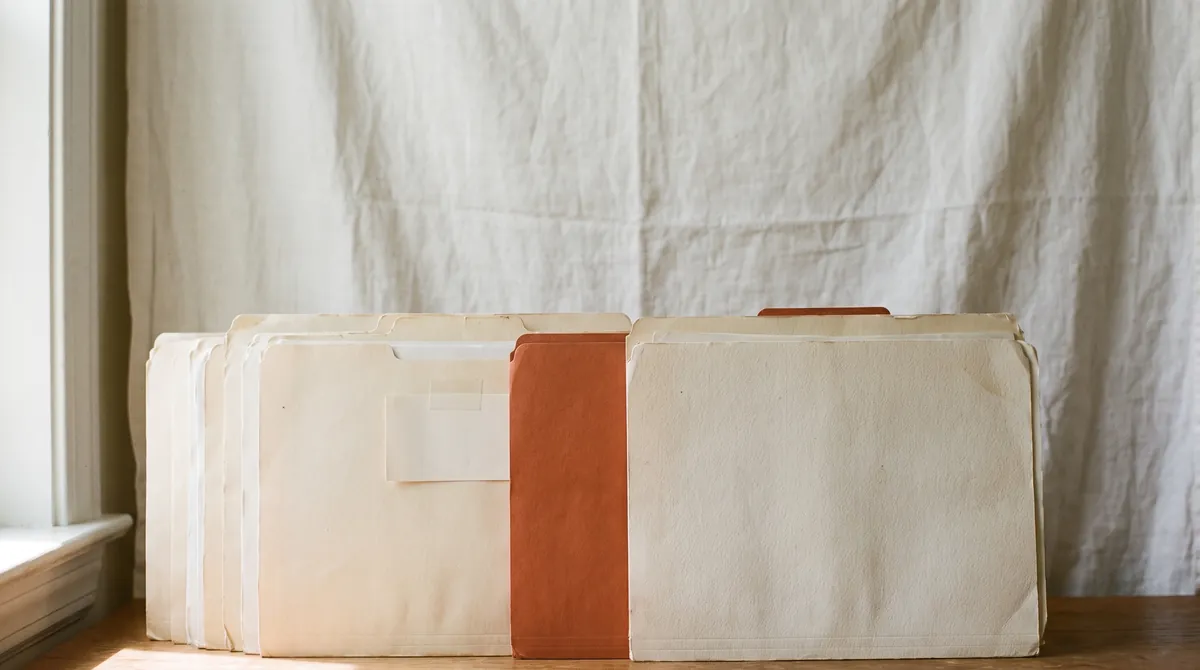 A row of worn cream file folders on a wooden desk with a single burnt orange folder standing among them